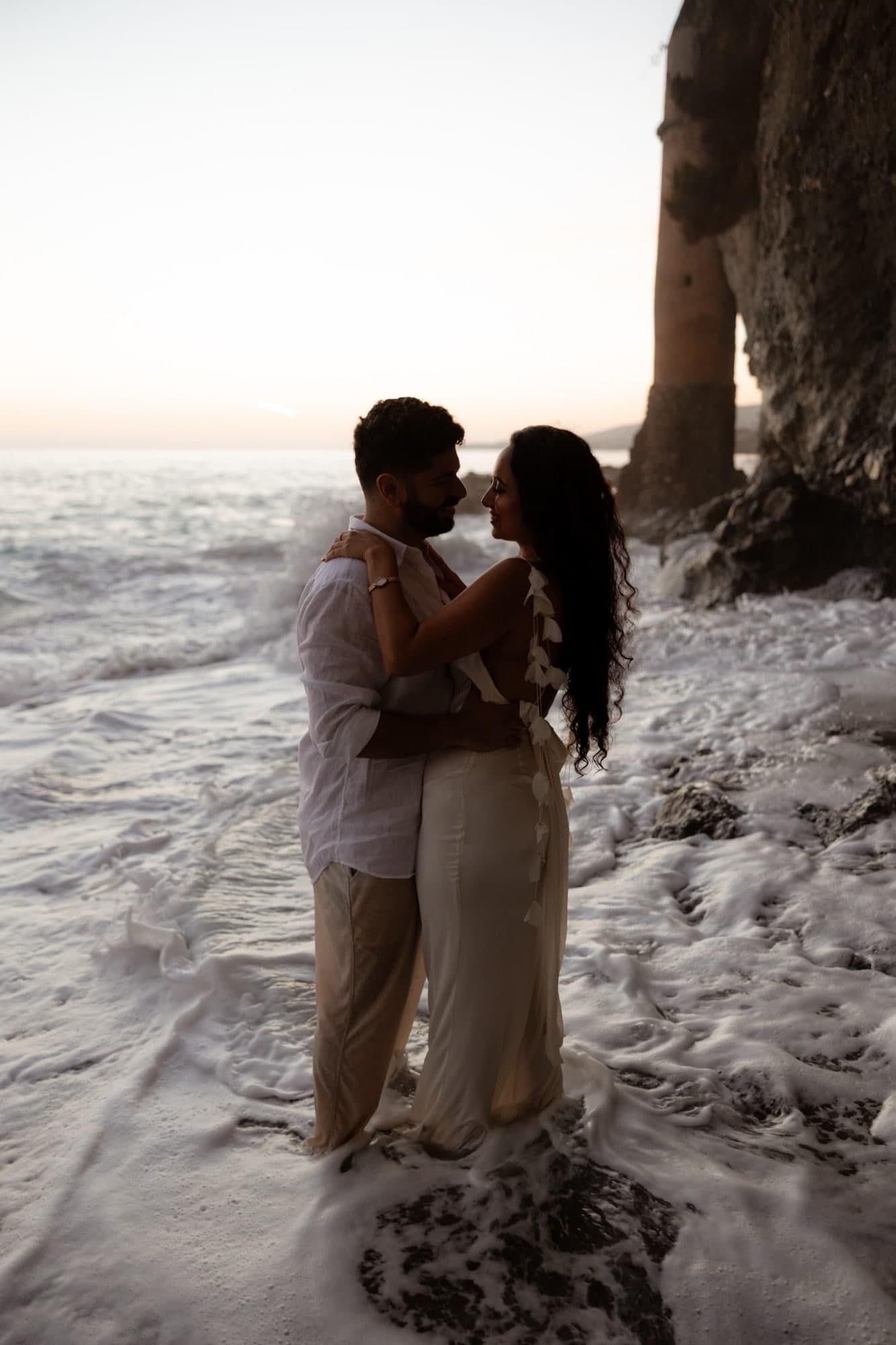 David and Joy embracing in the surf at sunset beside a stone sea tower