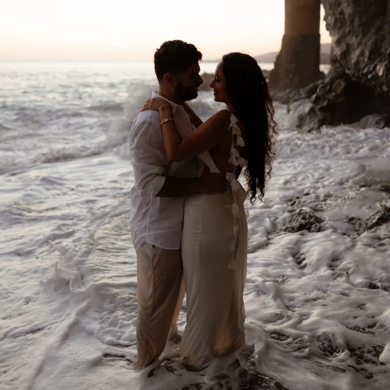 David and Joy embracing in the surf at sunset beside a stone sea tower